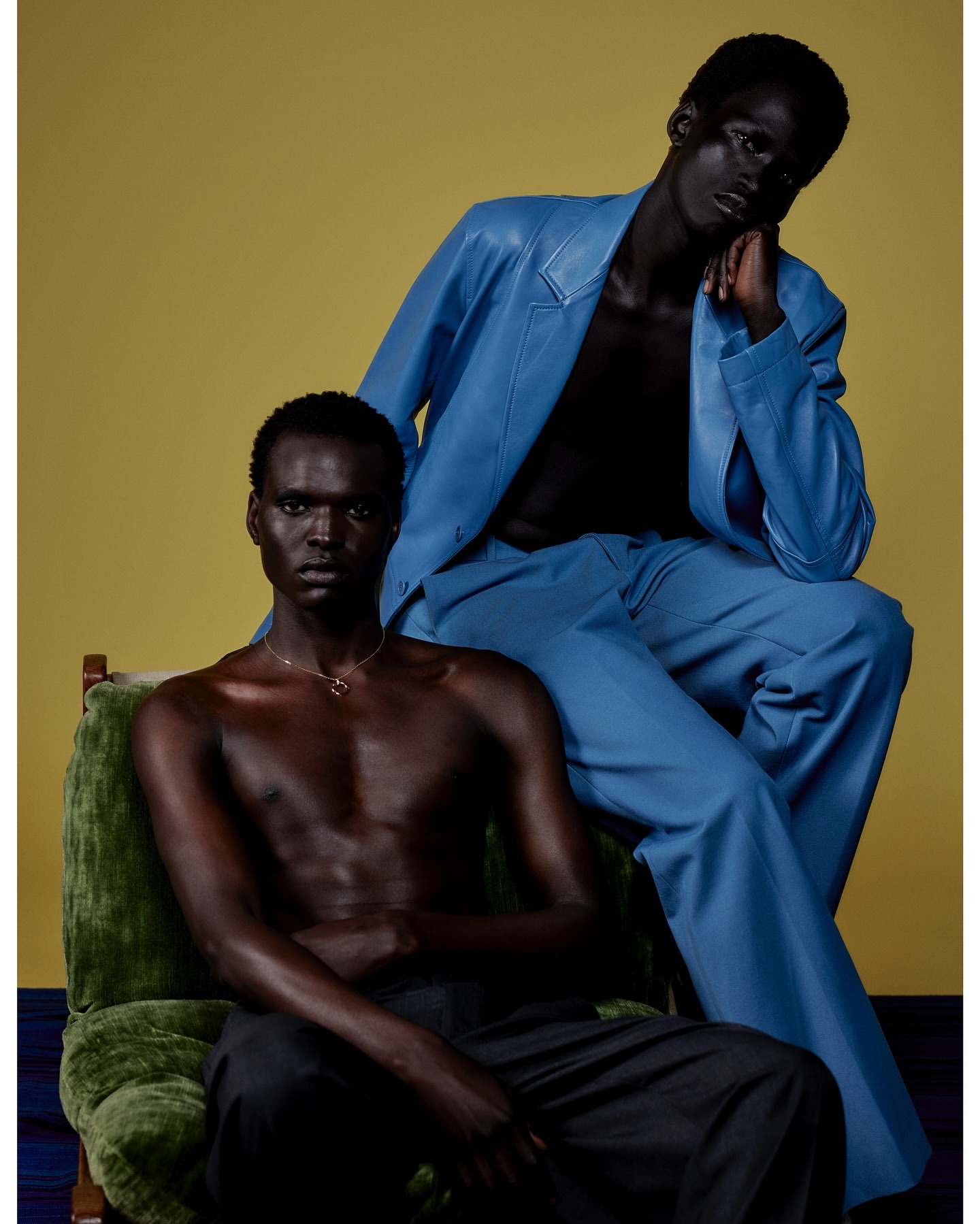 Two dark-skinned male fashion models in stylish clothing posing in a studio with a mustard-yellow background and blue flooring.
