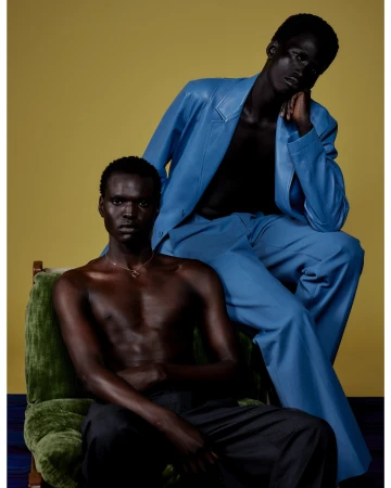 Two dark-skinned male fashion models in stylish clothing posing in a studio with a mustard-yellow background and blue flooring.