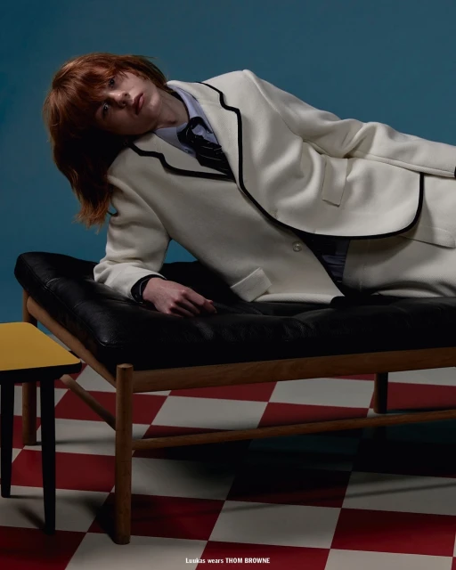 Fashion editorial shot of a redhead adult posing on a black chaise lounge wearing a cream-colored suit in a studio with a checkered floor.