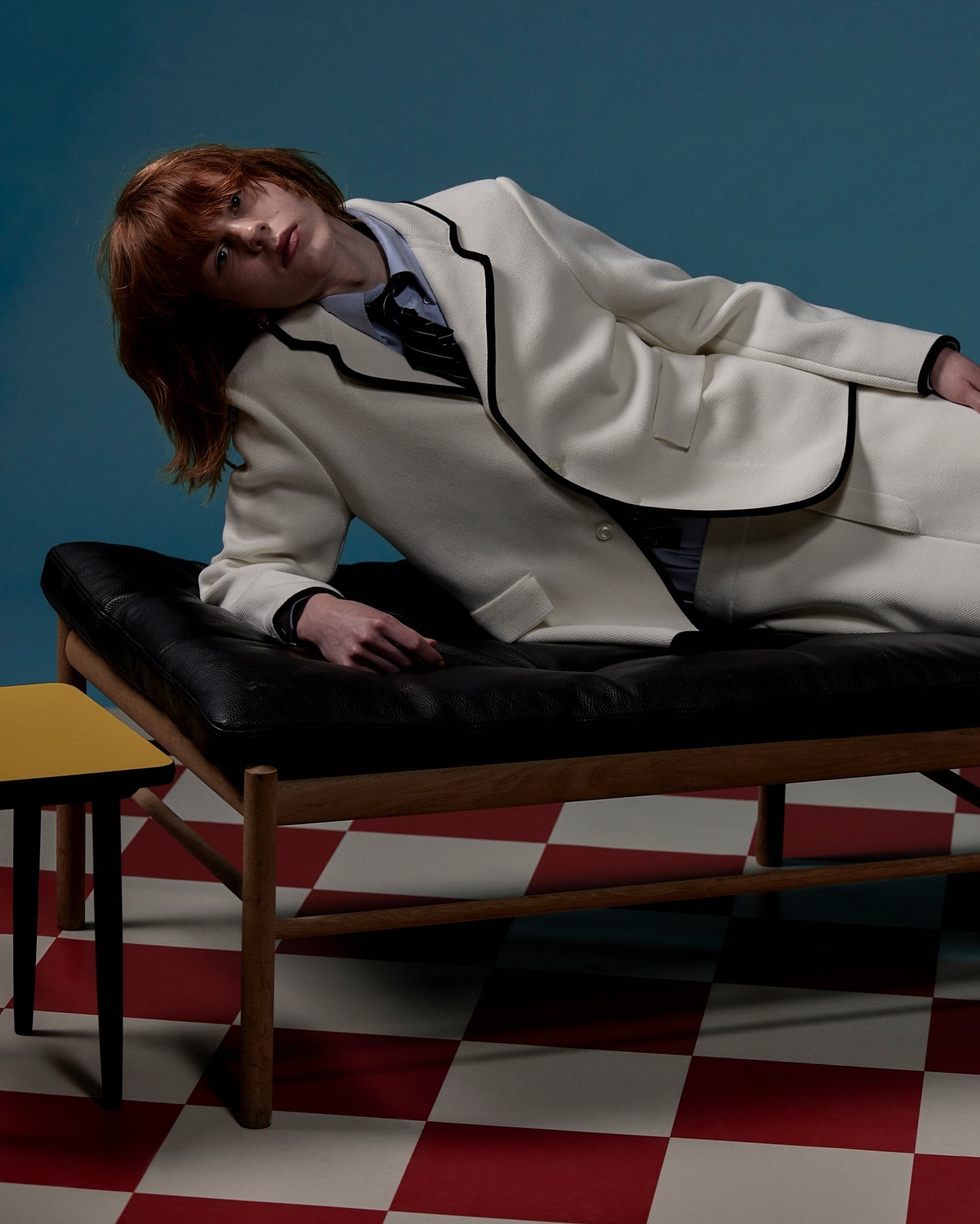 Adult laying on a black leather chaise lounge in white suit in a photo studio with a blue backdrop and red and white checkered floor.