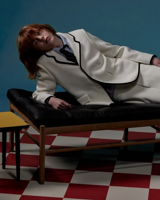 Adult laying on a black leather chaise lounge in white suit in a photo studio with a blue backdrop and red and white checkered floor.