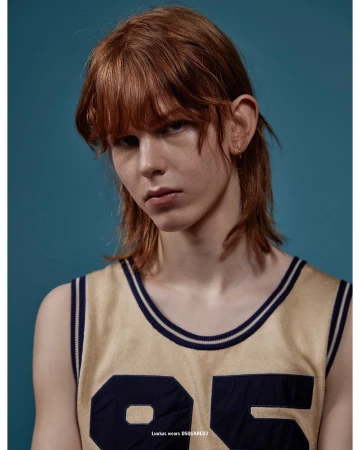 Young adult model posing indoors in a sleeveless shirt with the number 95. Close-up shot, light skin, auburn hair, teal background.