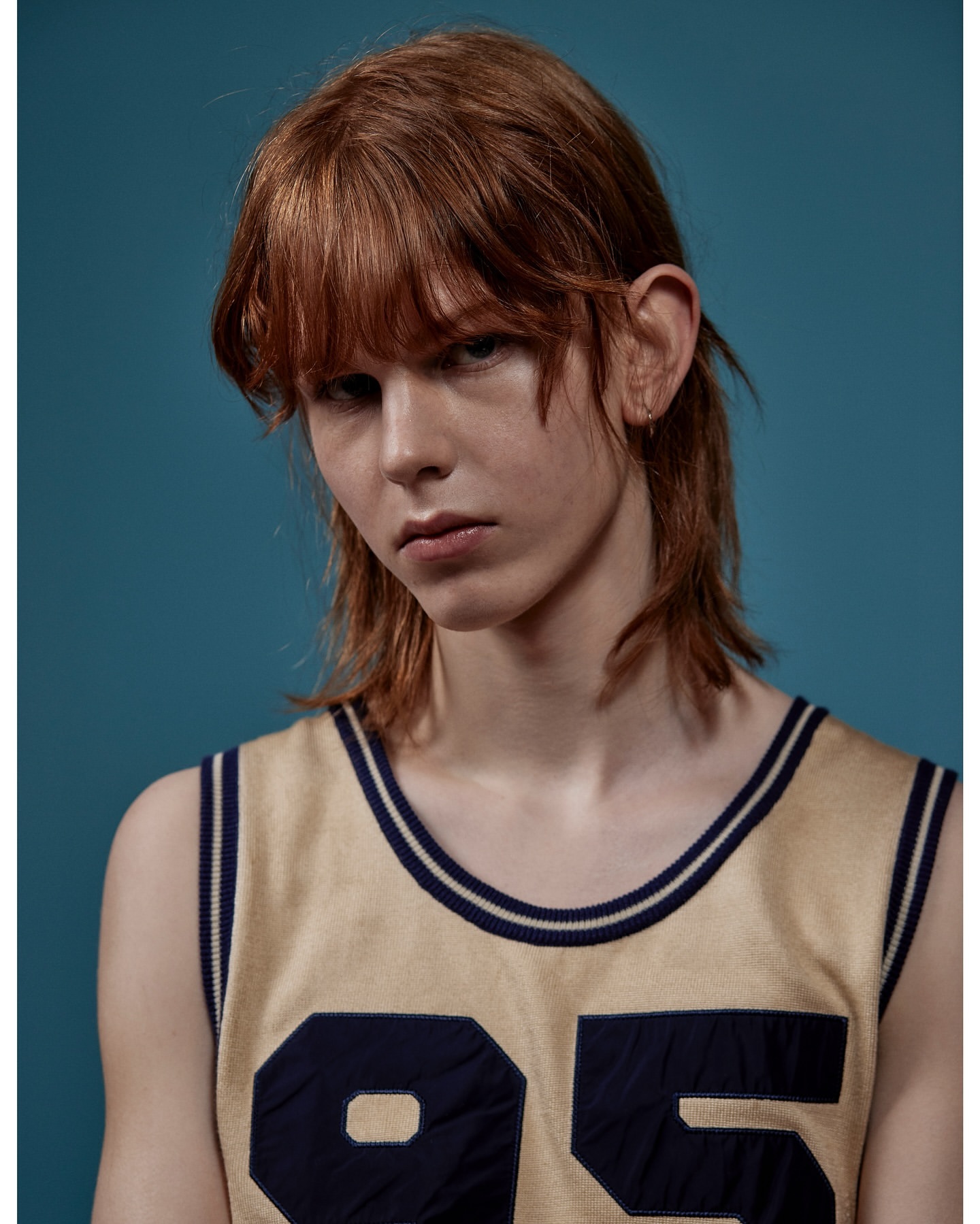 Portrait of a young adult with light skin and reddish-brown hair, wearing a sleeveless jersey with the number 95 against a solid blue backdrop.