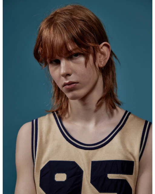 Portrait of a young adult with light skin and reddish-brown hair, wearing a sleeveless jersey with the number 95 against a solid blue backdrop.