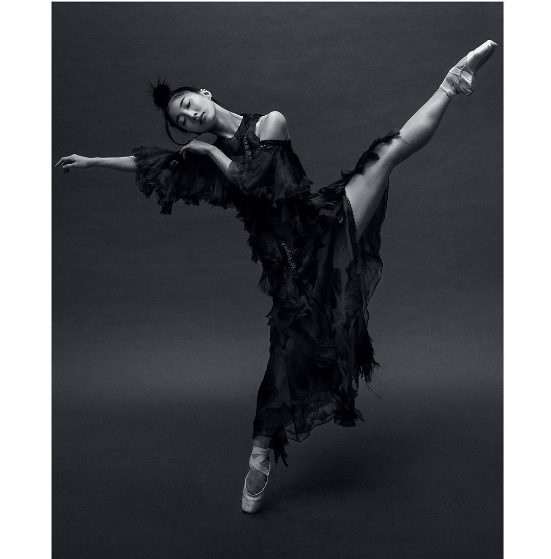 Female ballet dancer in pointe shoes poses in a dramatic full split wearing a long flowing black dress against a dark grey backdrop.