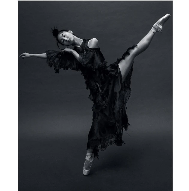 Female ballet dancer in pointe shoes poses in a dramatic full split wearing a long flowing black dress against a dark grey backdrop.