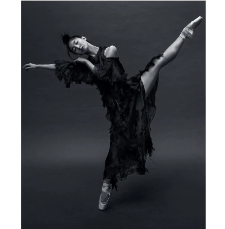 Female ballet dancer in pointe shoes poses in a dramatic full split wearing a long flowing black dress against a dark grey backdrop.