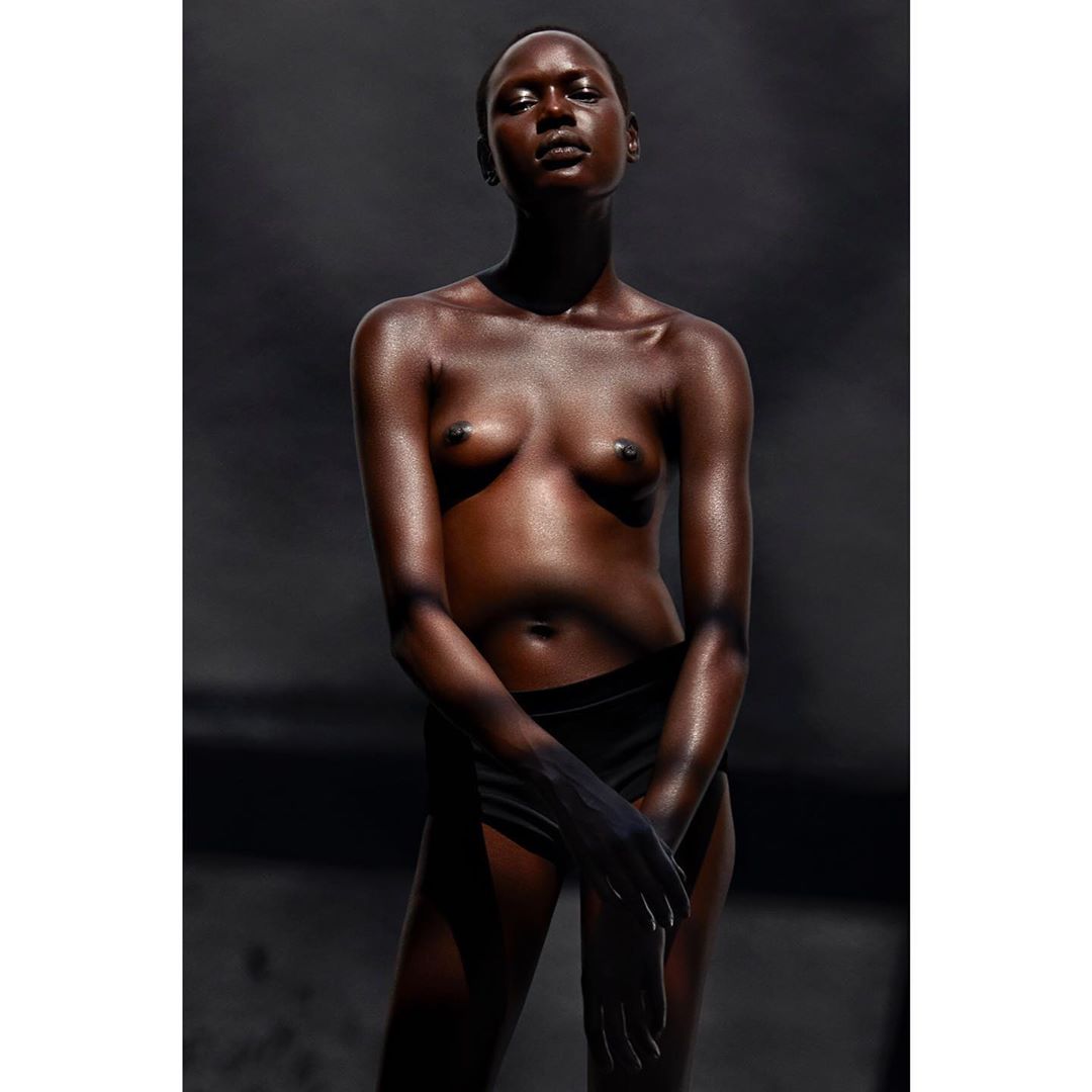 Artistic portrait of a nude adult woman with dark skin standing in a studio with her hands together and dark background.