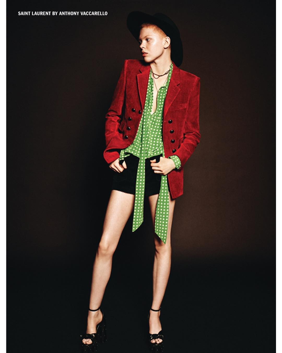 A fair-skinned fashion model in a red corduroy jacket, green polka dot blouse, and black shorts poses against a dark brown backdrop.