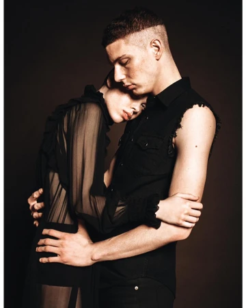 Studio shot of a man and woman in a close embrace, both in black clothing, against a dark background, conveying a sense of intimacy and fashion.