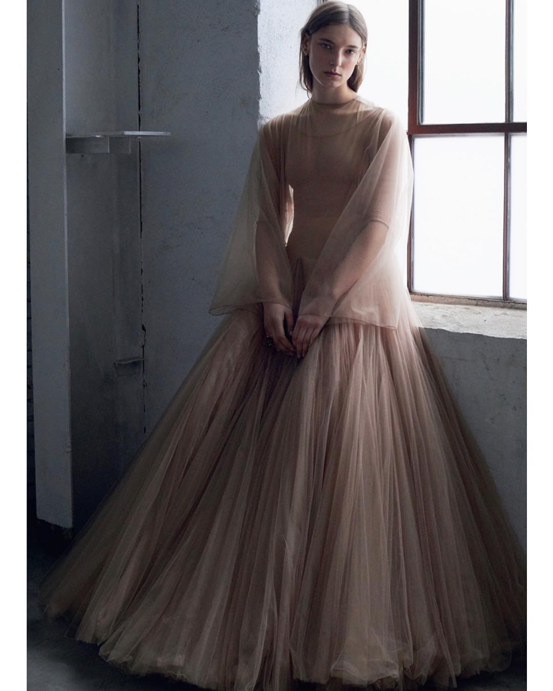 Elegant woman in flowing, layered gown sits near a window in a muted, neutral color palette, photographed in a studio setting.