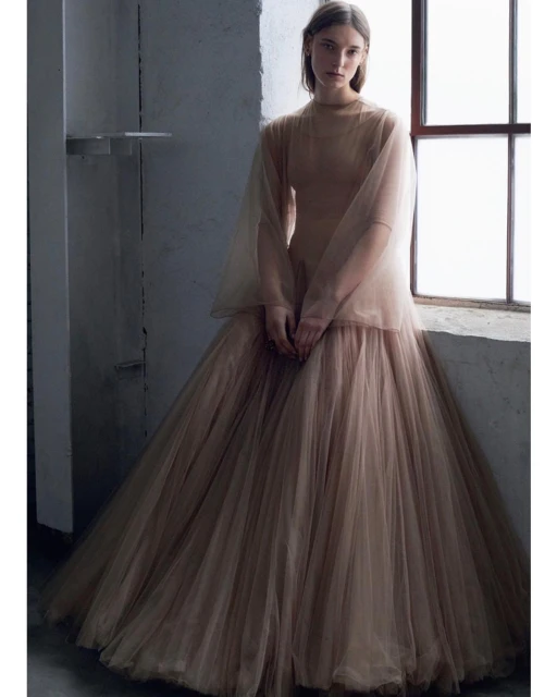Elegant woman in flowing, layered gown sits near a window in a muted, neutral color palette, photographed in a studio setting.