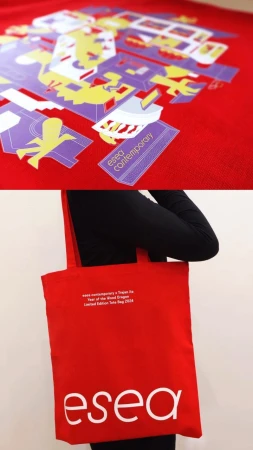 A red tote bag with the logo 'esea' in white is held by a person wearing black, with a colorful design on a red background above.