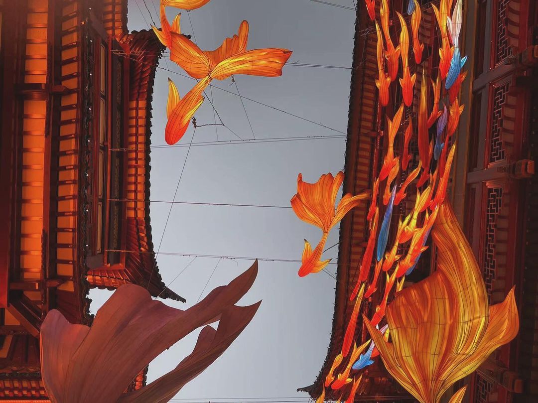 Orange fish-shaped lanterns hang over a street in an urban area, with traditional architecture visible on either side under an overcast sky.