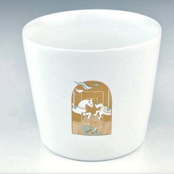 Close-up of a white ceramic cup with a carousel horse illustration in gold and blue hues, set against a neutral studio backdrop.