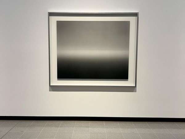 Black and gray abstract art print hangs on a white wall above a gray tiled floor in a modern, minimalist art gallery or museum.