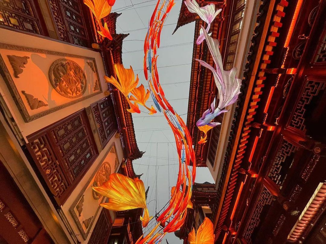 Whimsical street scene featuring colorful decorative fish hanging between traditional Chinese buildings with intricate architectural details.
