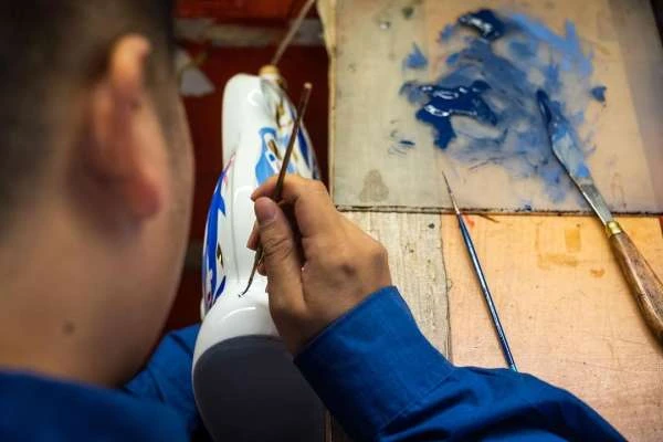 Artist meticulously paints blue and white pattern on ceramic vase with a fine brush indoors; art supplies and tools visible.
