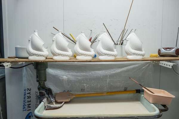 Still life with white ceramic ship sculptures displayed on a shelf with paint brushes and tools, set against a white wall.