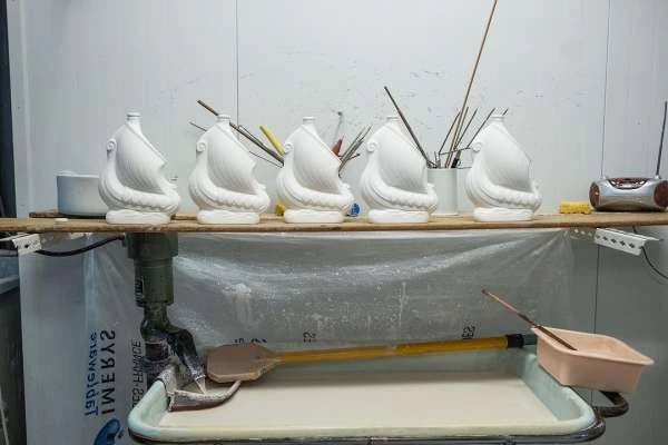 Still life with white ceramic ship sculptures displayed on a shelf with paint brushes and tools, set against a white wall.