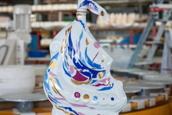 Close-up of a decorative white ceramic ship with blue, pink, and gold details inside a factory setting with blurred machinery.
