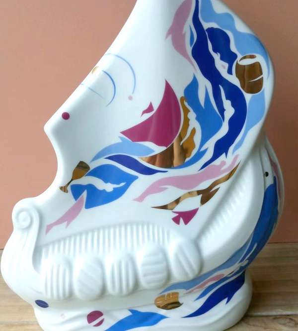 Close-up of a white ceramic vessel with painted blue, pink, and gold abstract designs, evoking nautical and maritime themes on a wooden surface.