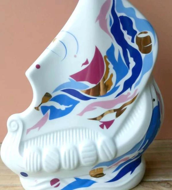 Close-up of a white ceramic vessel with painted blue, pink, and gold abstract designs, evoking nautical and maritime themes on a wooden surface.