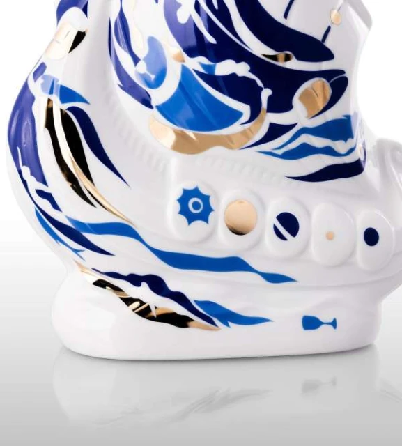 Ceramic ship figurine in white, blue, black, and gold on a white reflective surface. Ornamental sea designs and gold detailing.
