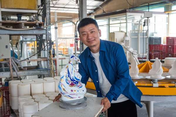 Adult man in a blue button-down shirt next to a decorative ceramic swan-like object in an industrial setting with tables and equipment.