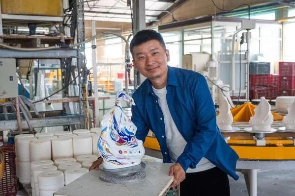 Adult man in a blue button-down shirt next to a decorative ceramic swan-like object in an industrial setting with tables and equipment.