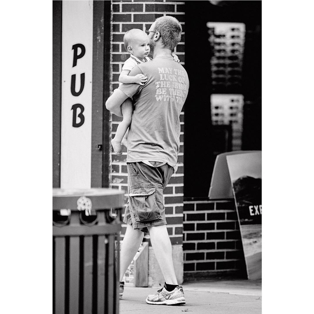 Black-and-white shot of dad carrying a baby in an urban environment, with building brick walls as backdrop, creating a candid, family moment.