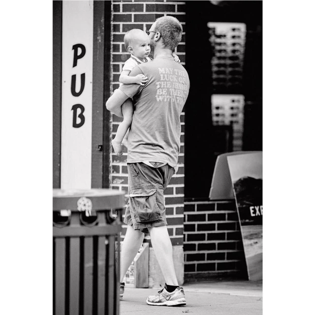 Black-and-white shot of dad carrying a baby in an urban environment, with building brick walls as backdrop, creating a candid, family moment.