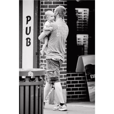 Black-and-white shot of dad carrying a baby in an urban environment, with building brick walls as backdrop, creating a candid, family moment.