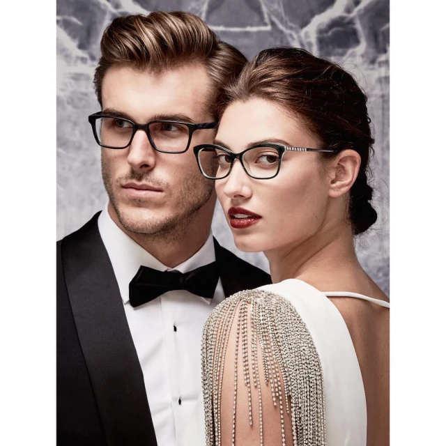 A stylish couple poses indoors; the man wears a tuxedo and glasses, and the woman is in a white gown with glasses and fringe details.