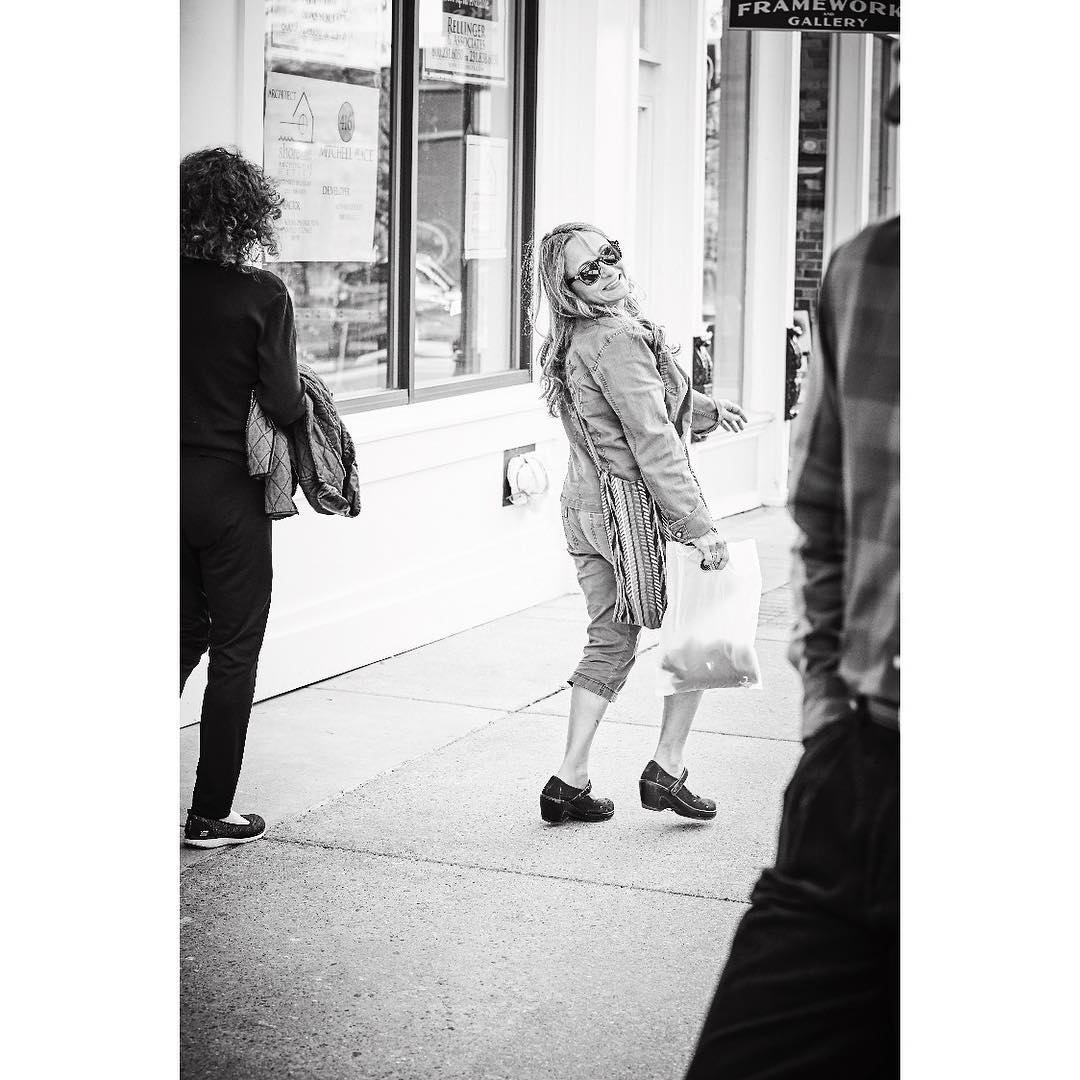 Black and white street scene featuring a woman in a denim jacket and sunglasses looking back with a shopping bag, walking on the sidewalk.