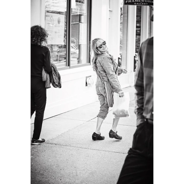 Black and white street scene featuring a woman in a denim jacket and sunglasses looking back with a shopping bag, walking on the sidewalk.