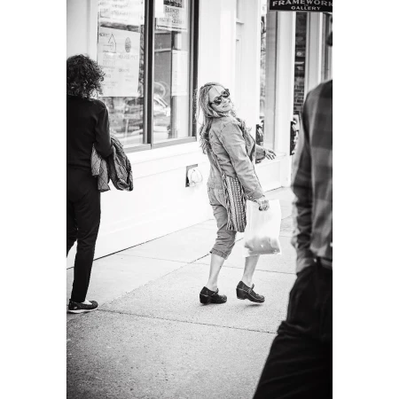 Black and white street scene featuring a woman in a denim jacket and sunglasses looking back with a shopping bag, walking on the sidewalk.