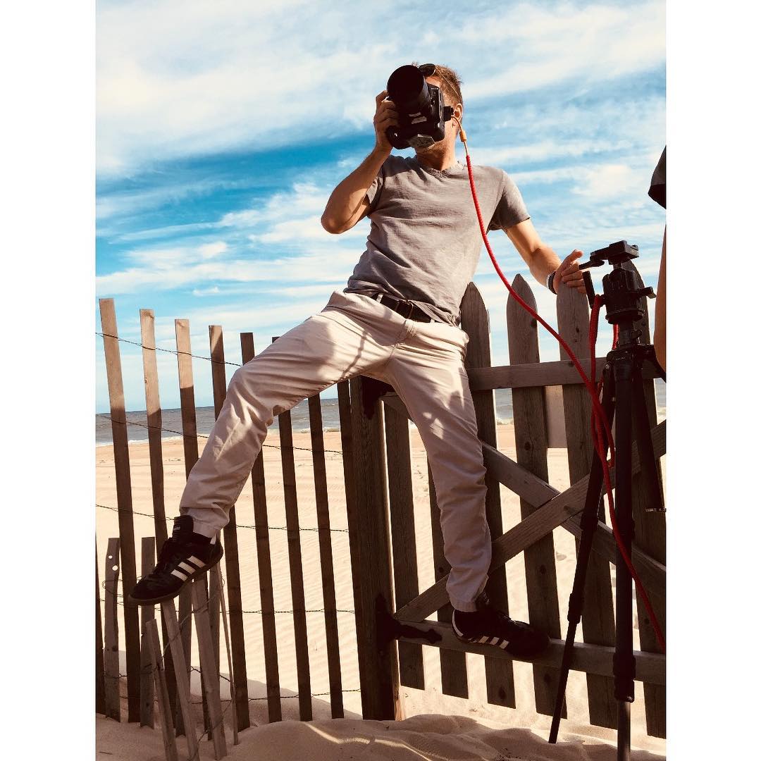 A photographer stands on a wooden fence at the beach, holding a camera up to his face, taking a photo, on a partly cloudy day.