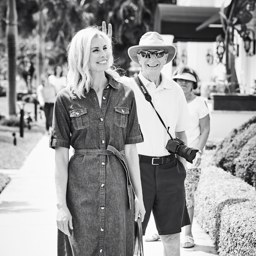 A woman in a denim dress and an older man wearing a hat and holding a camera walk side-by-side outdoors on a sunny day.