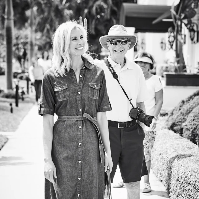 A woman in a denim dress and an older man wearing a hat and holding a camera walk side-by-side outdoors on a sunny day.