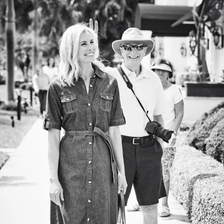 A woman in a denim dress and an older man wearing a hat and holding a camera walk side-by-side outdoors on a sunny day.