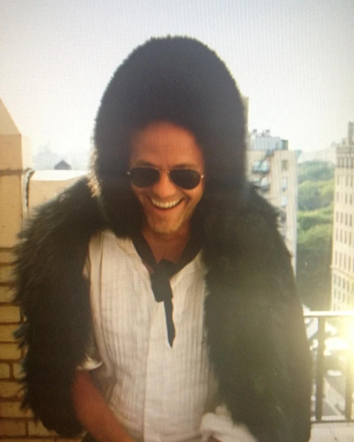 Smiling man outdoors wearing sunglasses and a large black fur hood and white shirt in an urban outdoor environment with buildings.
