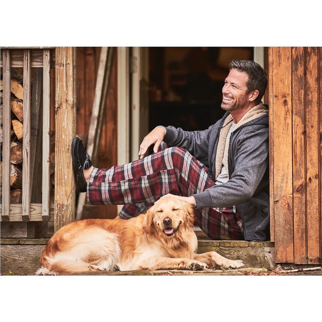 A man and a dog are sitting together on a porch with wooden posts and beams; firewood is stacked behind him, and he is smiling.