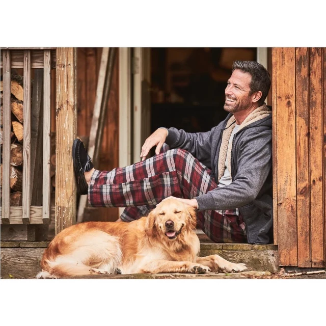 A man and a dog are sitting together on a porch with wooden posts and beams; firewood is stacked behind him, and he is smiling.
