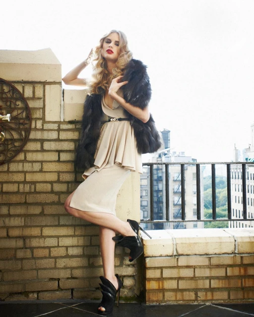 A fashionable woman in a beige dress and fur shawl poses on a rooftop with a city view, highlighting an elegant and urban aesthetic.