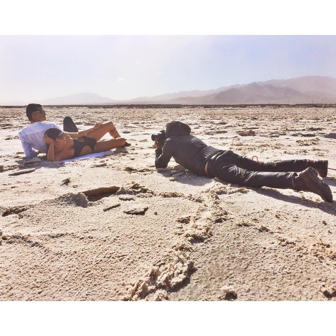 Fashion shoot on a dry lakebed with two adults posing in swimwear for a prone photographer in an arid landscape with mountains.