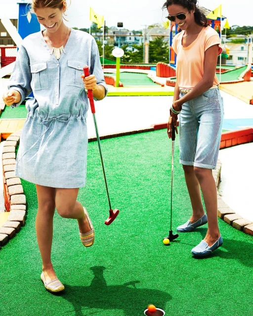 Two women smile while playing miniature golf on a green outdoor course with houses and trees visible in the distance on a sunny day.