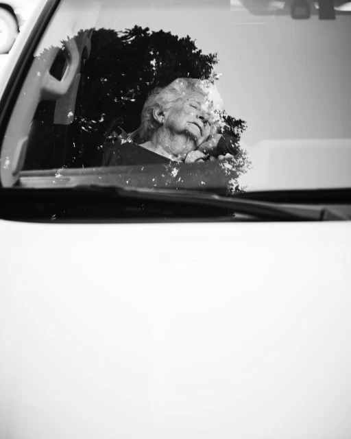 Black and white close-up of an elderly woman sleeping in the driver's seat of a car, her face partially obscured by tree reflections.