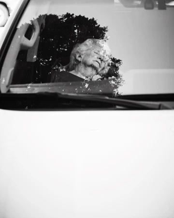 Black and white close-up of an elderly woman sleeping in the driver's seat of a car, her face partially obscured by tree reflections.