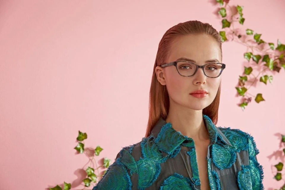Close-up of a woman with red hair wearing glasses and a green patterned jacket, posing in front of a pink background with green vines.